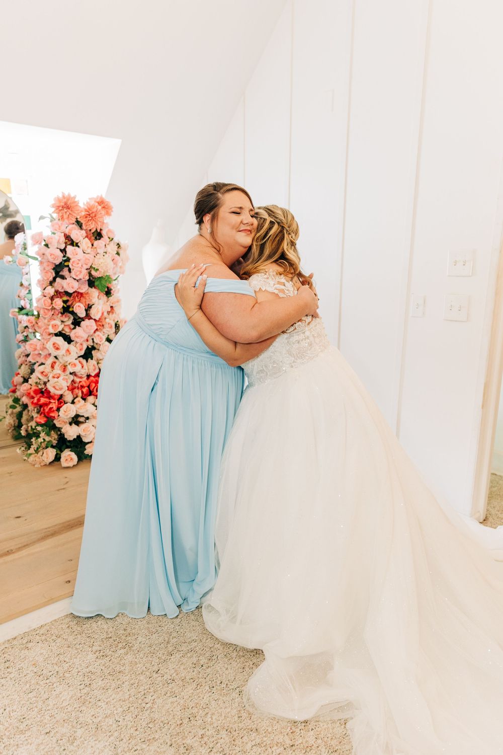 Emotional wedding scene with bridesmaids in light blue dresses hugging bride in white gown near floral arrangements.