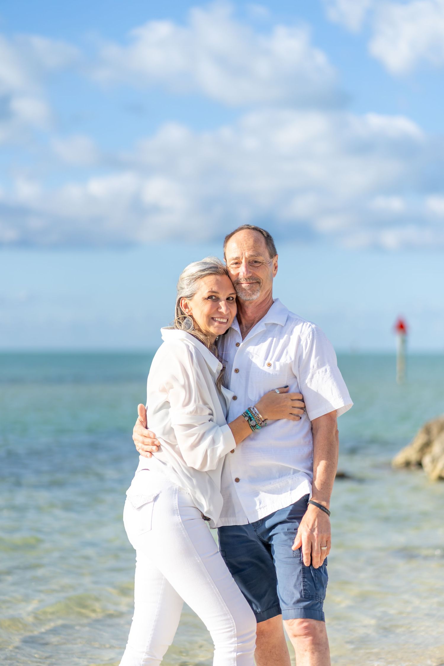 A couple in white outfits walking along a sunny tropical beach near the water.