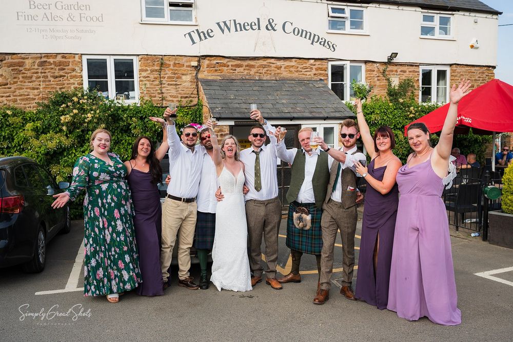The wedding party poses outside The Wheel and Compass pub with guests in purple and white formal attire.