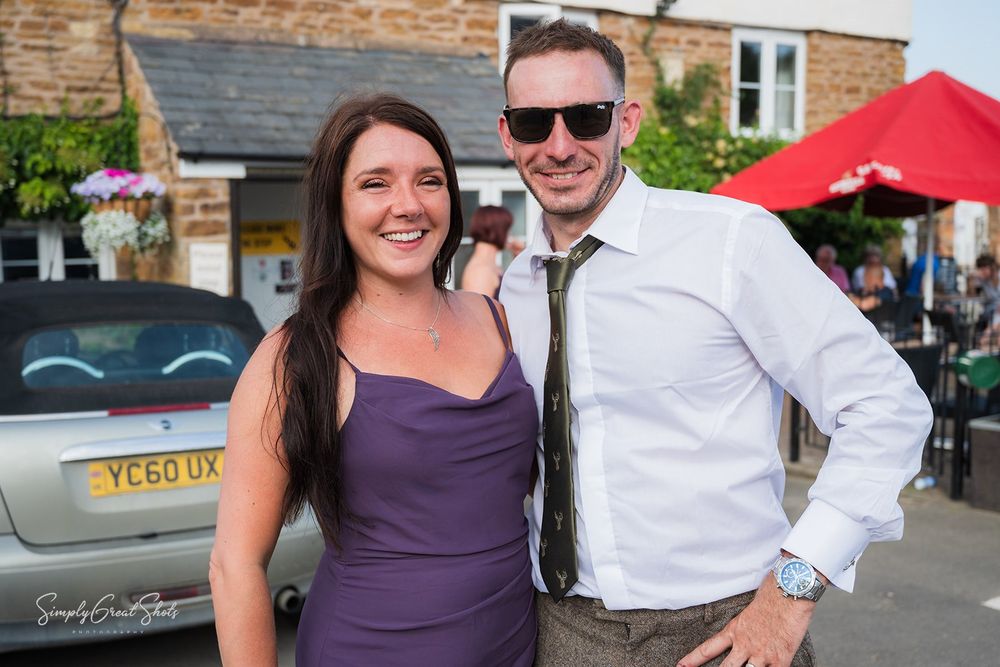 Two people in formal wedding attire smile and pose together outside the Wheel and Compass public house on a sunny day.
