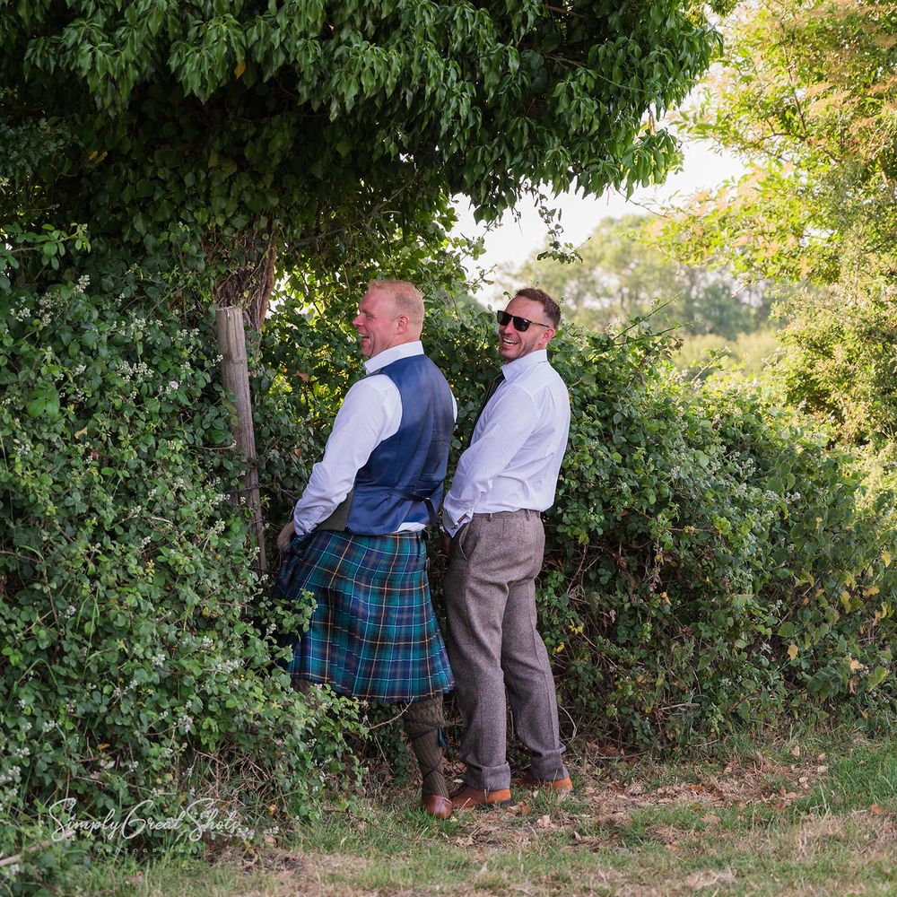 The groom and best man are standing in front of trees answering a call of nature, in an outdoor nature setting during daytime.
