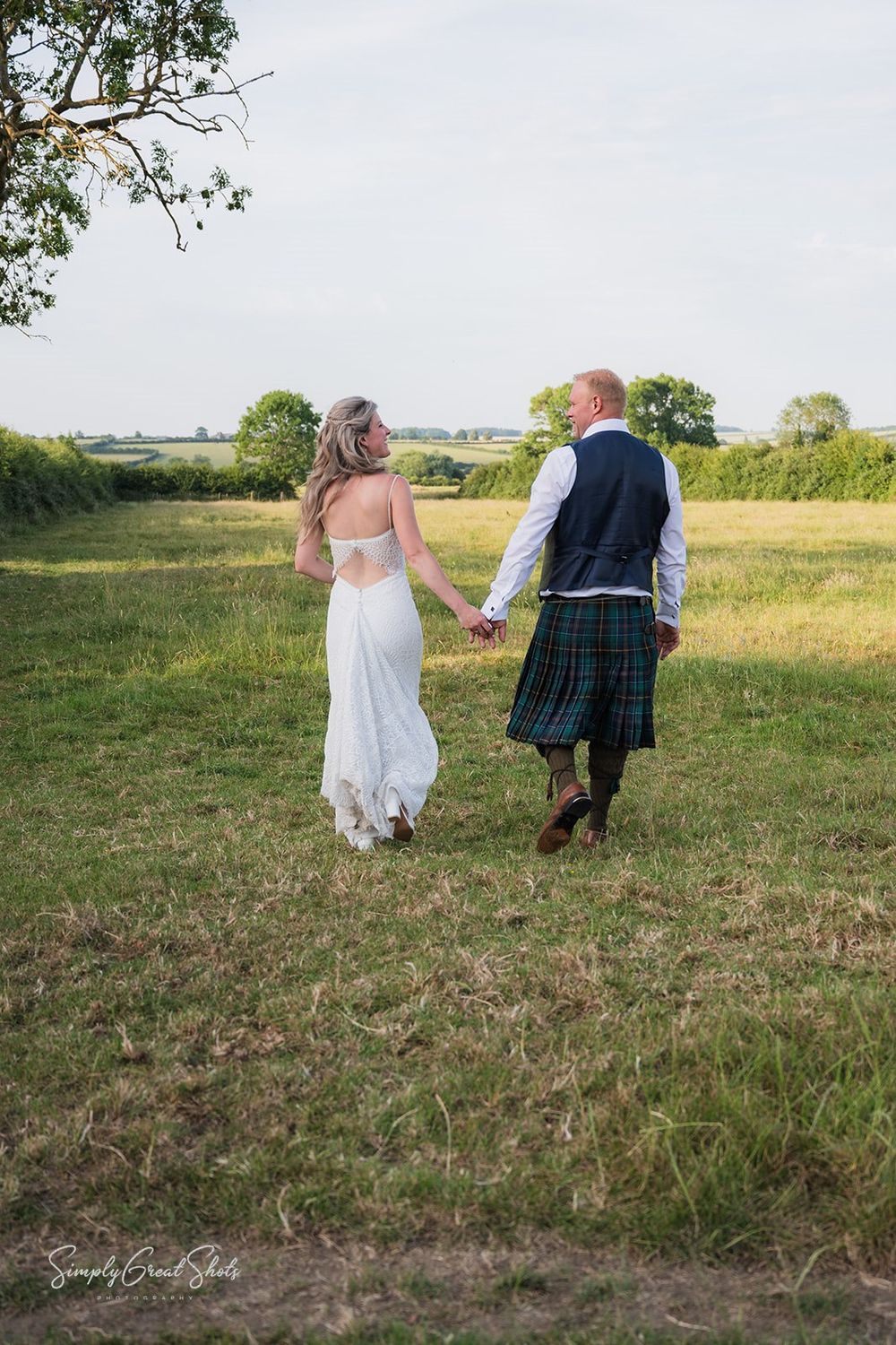 A happily married couple holding hands walks through a grassy rural field while wearing wedding attire and a traditional Scottish kilt.