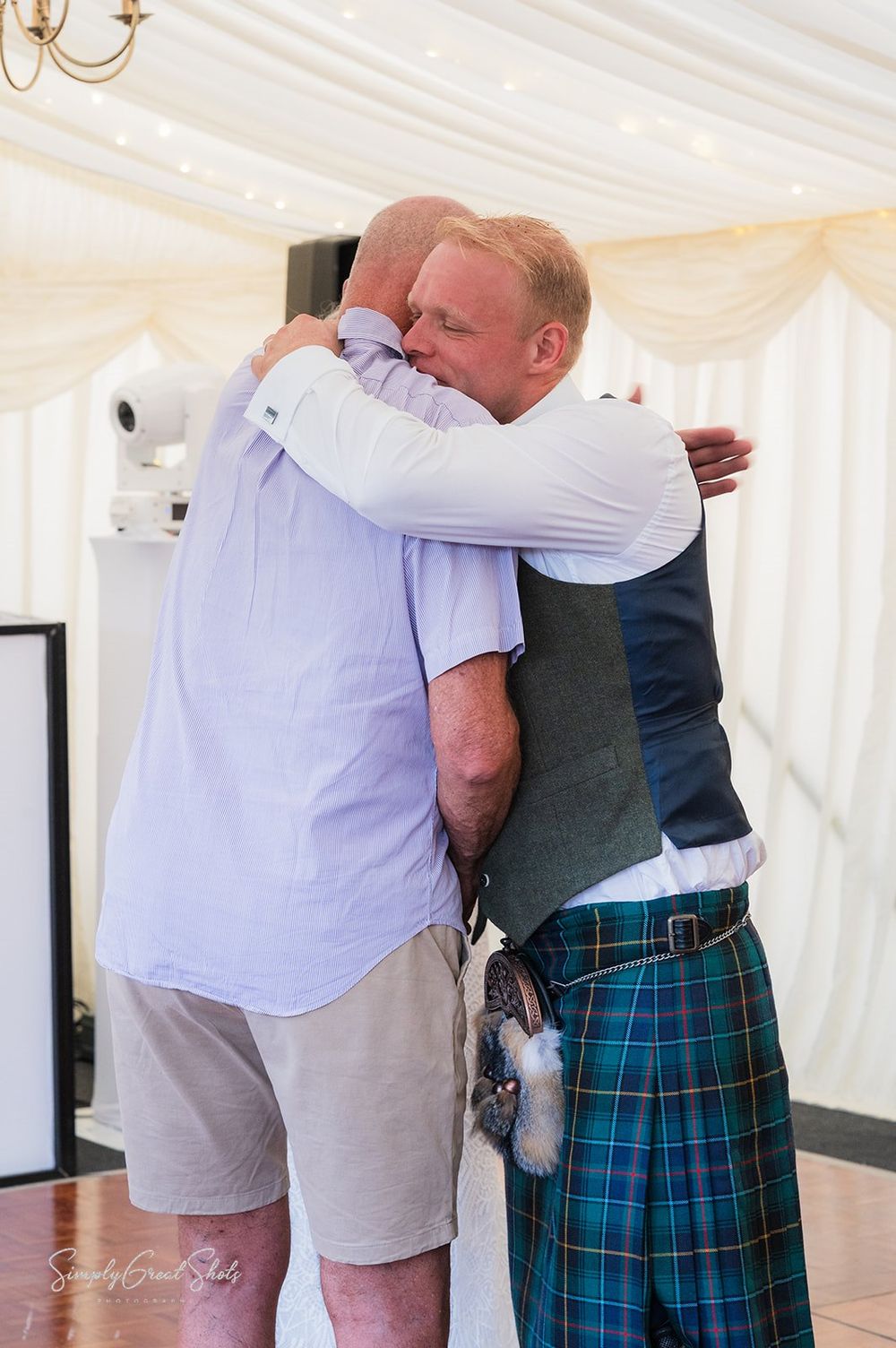 The father of the bride and groom share an emotional embrace at a wedding, with one wearing traditional Scottish tartan kilt attire.