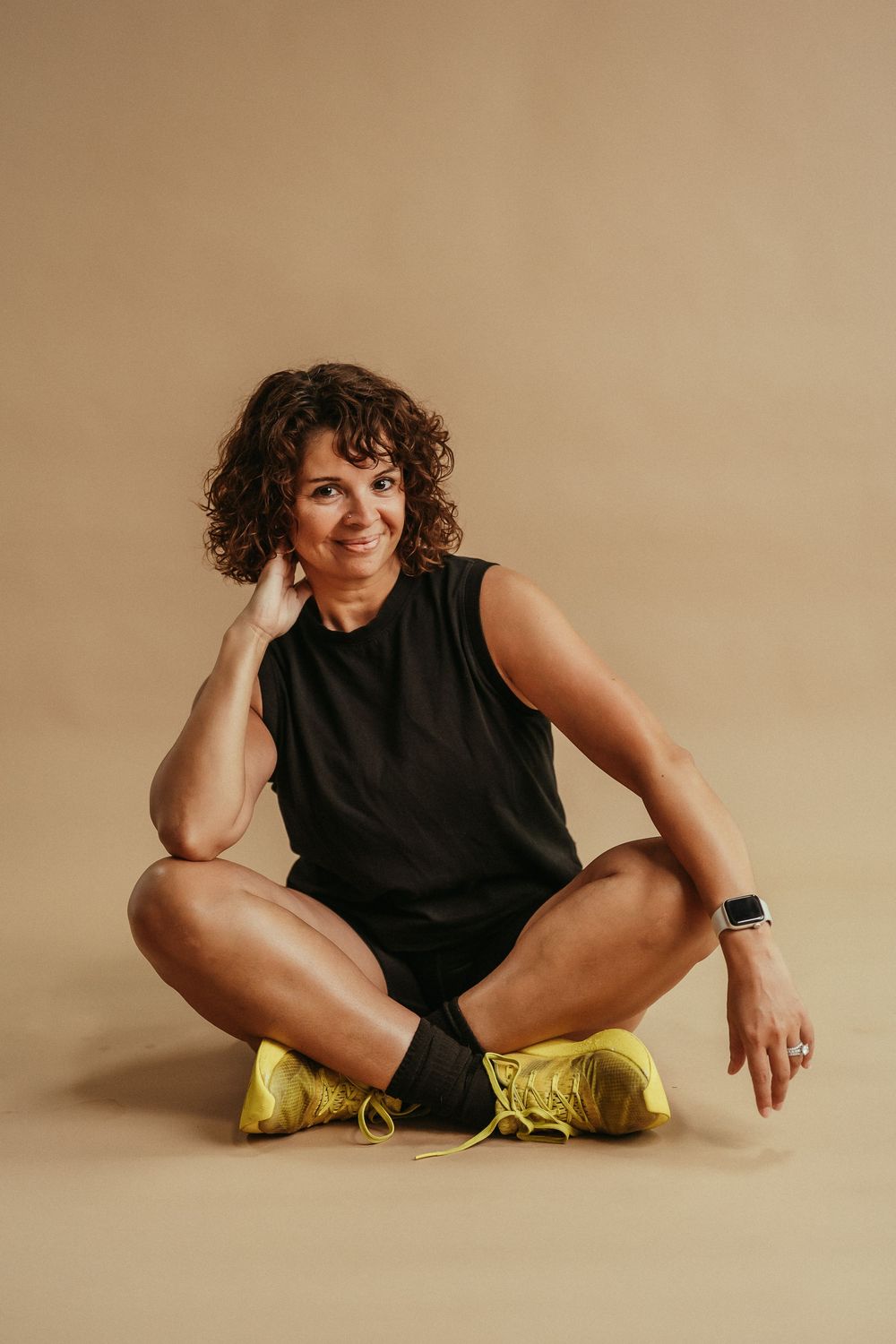 Athletic person in black sleeveless top and shorts sits cross-legged on floor wearing bright yellow sneakers.