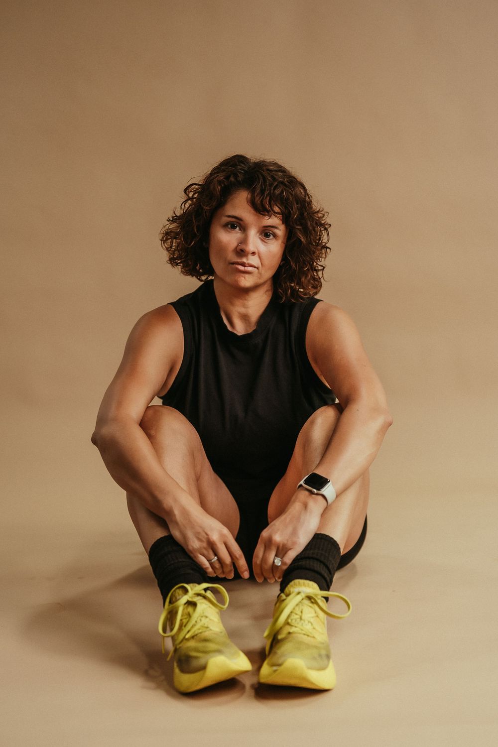 Athletic person in black tank top and yellow sneakers sits on beige background tying shoelaces.