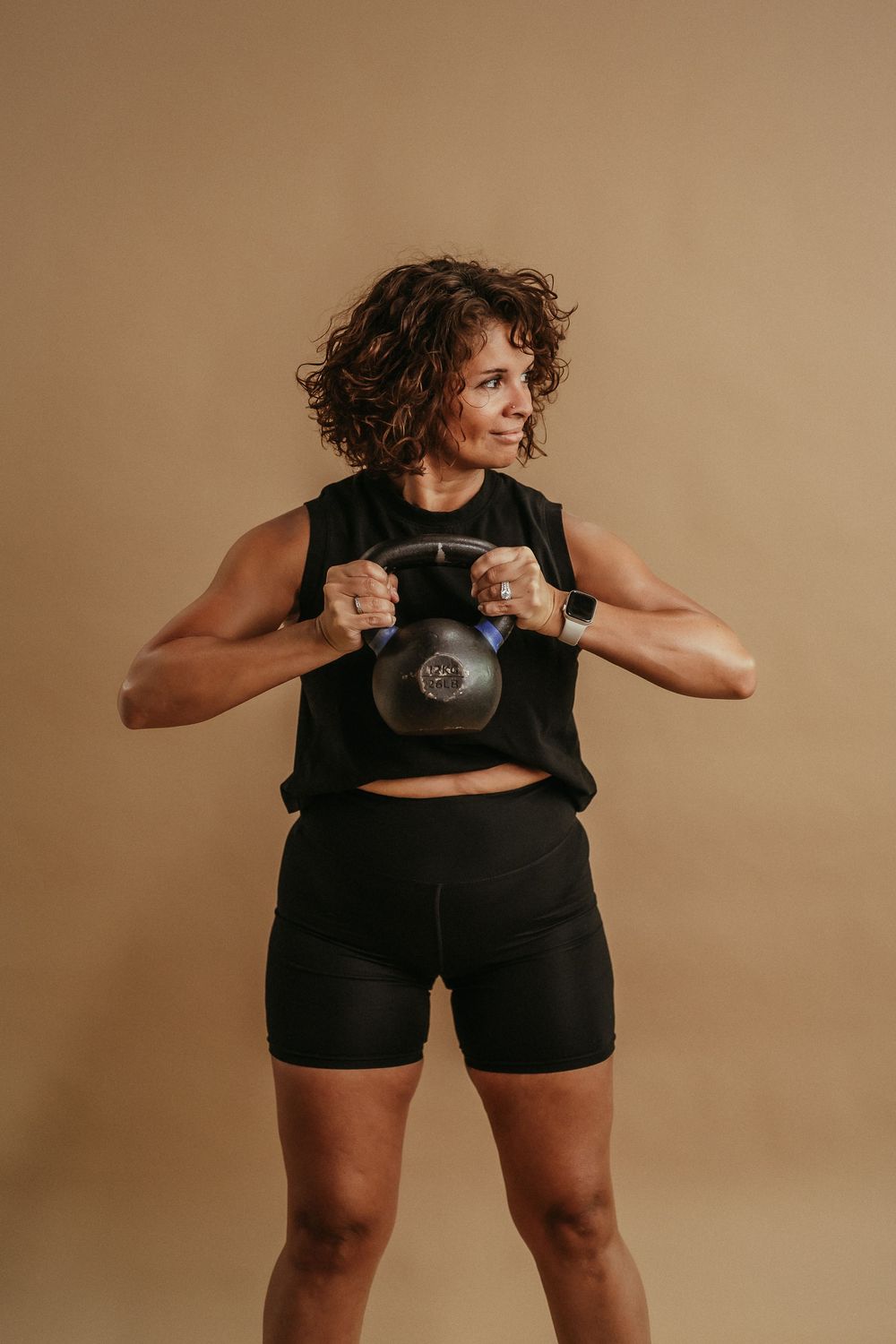 Person in workout attire demonstrates proper form while lifting a heavy kettlebell against beige backdrop.
