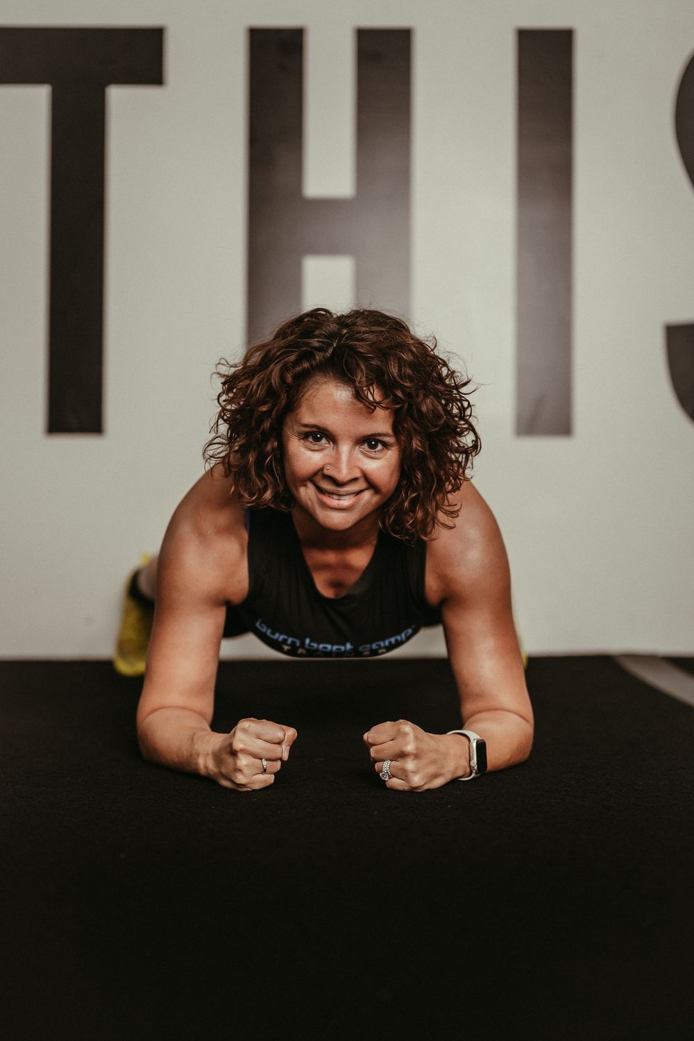 Athletic figure in black tank top smiling and doing workout poses against dark background.