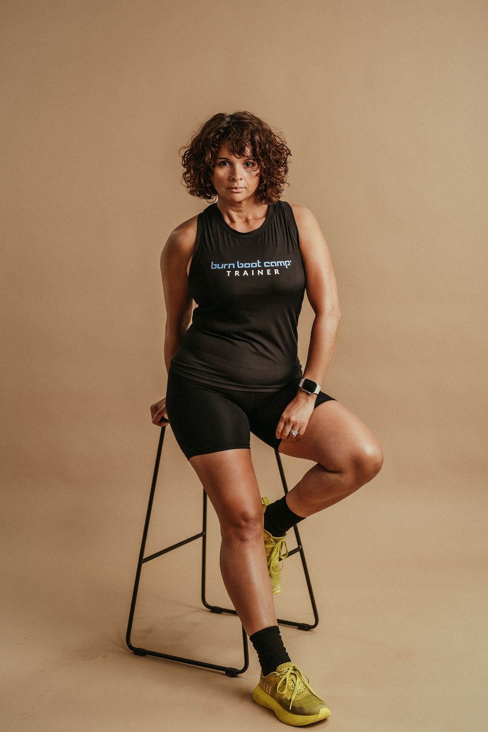 Person wearing black athletic tank top sitting on exercise chair with yellow sneakers against beige backdrop.