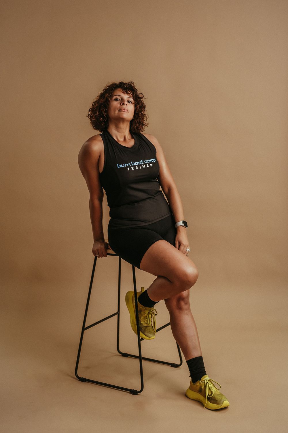 Athletic person in black workout attire sits on tall stool wearing bright yellow sneakers against beige studio backdrop.