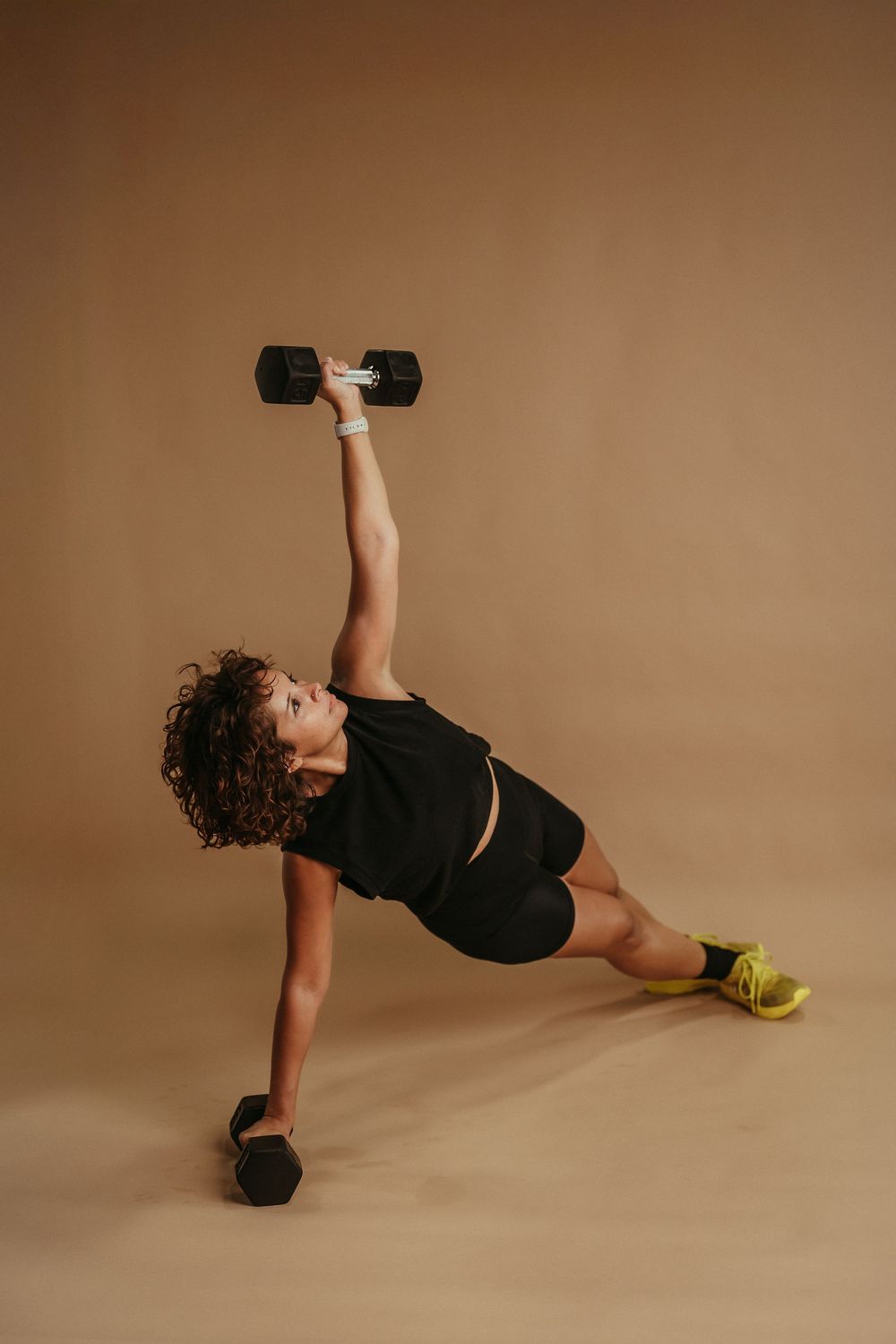 Athletic woman in black workout clothes performs a side plank exercise with dumbbells against brown backdrop.