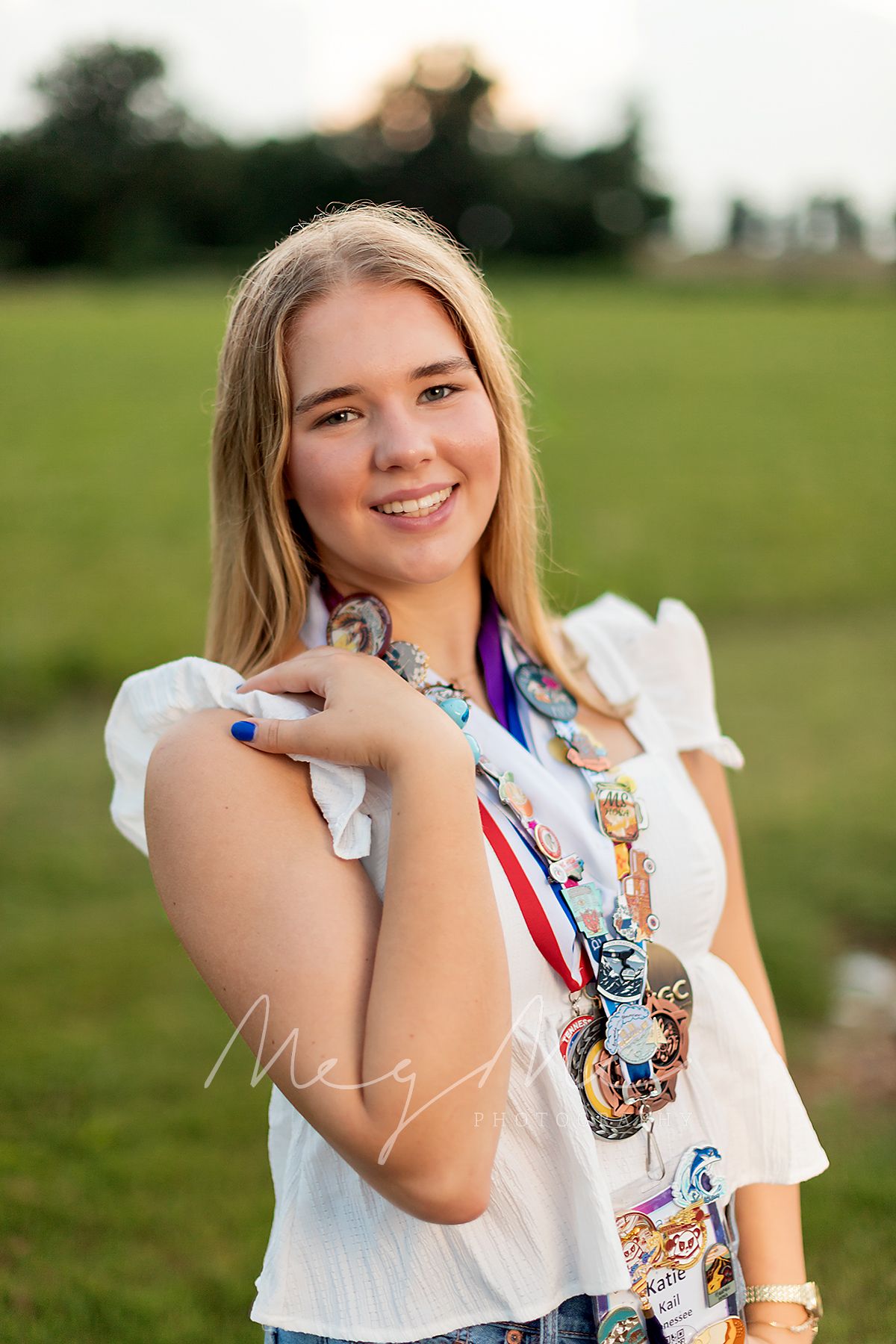 Teen girl in a field during golden hour, wearing a white blouse and decorated with medals. Senior Pictures by MegMc Photography, Medina, TN
