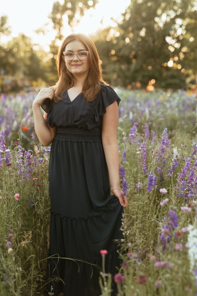 Person in a black dress stands in a meadow of purple and pink wildflowers during sunset.