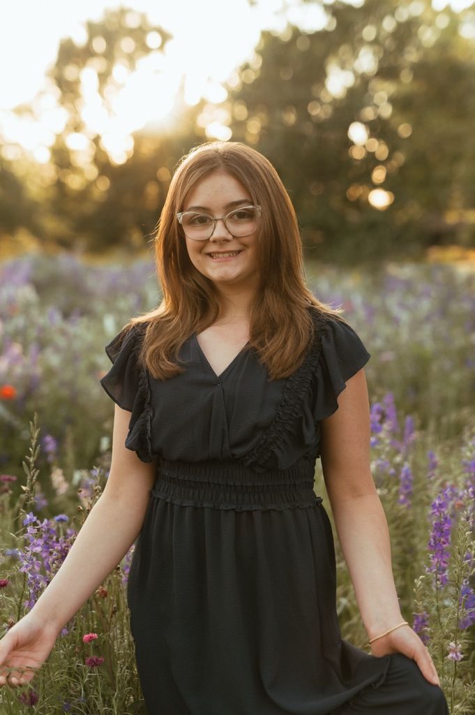 Woman wearing black dress and glasses standing in field of purple wildflowers at sunset.
