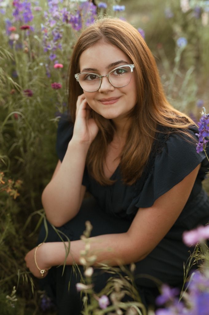 A portrait of someone wearing glasses and a black top posing among purple wildflowers during golden hour.