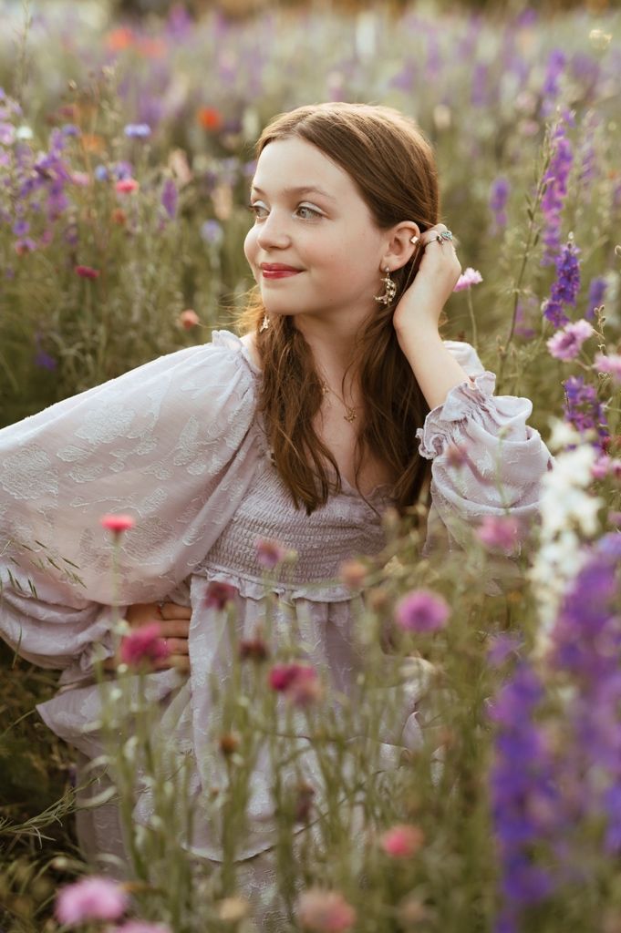 Young person in flowing white dress twirls gracefully among purple and pink wildflowers at sunset.