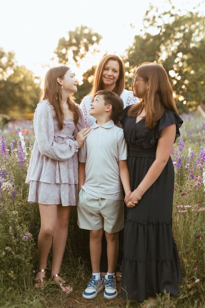 A family group shares a candid moment together while standing in a field of purple wildflowers at sunset.