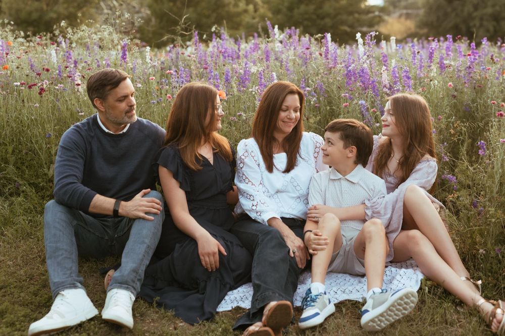 A family sits together on the ground laughing and bonding in a field with purple flowers during sunset.