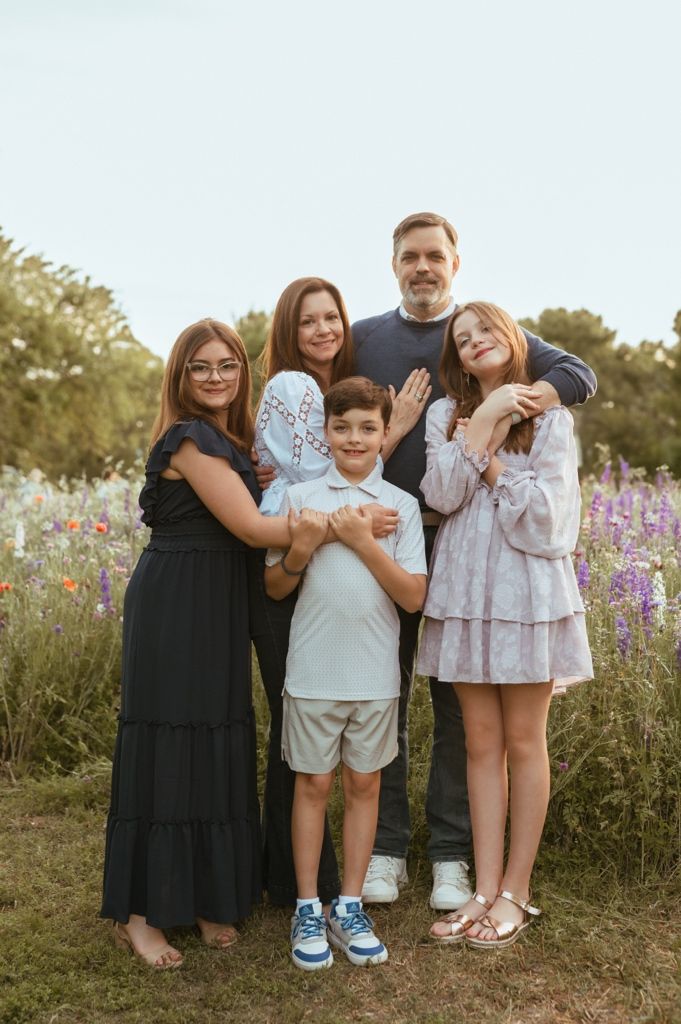 Family standing together outdoors in a wildflower meadow with purple and orange blooms at sunset.