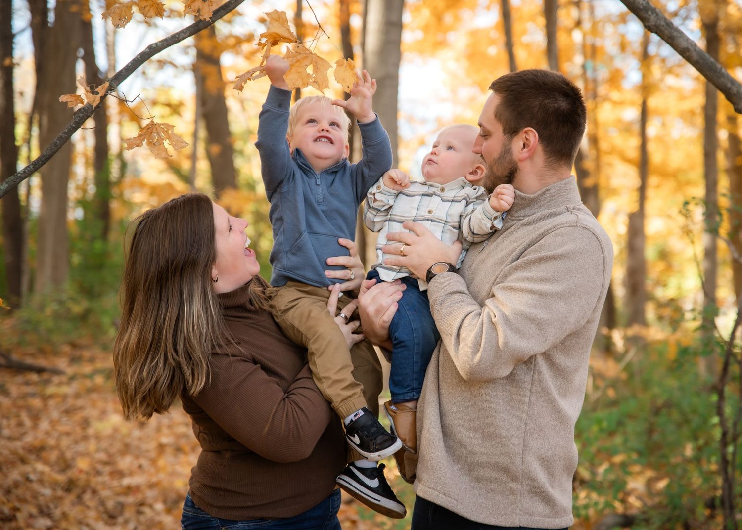 A joyful family moment captured outdoors during autumn with fallen leaves and golden trees in the background.