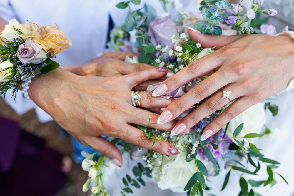 Hands adorned with rings resting on a wedding bouquet with purple and white flowers.