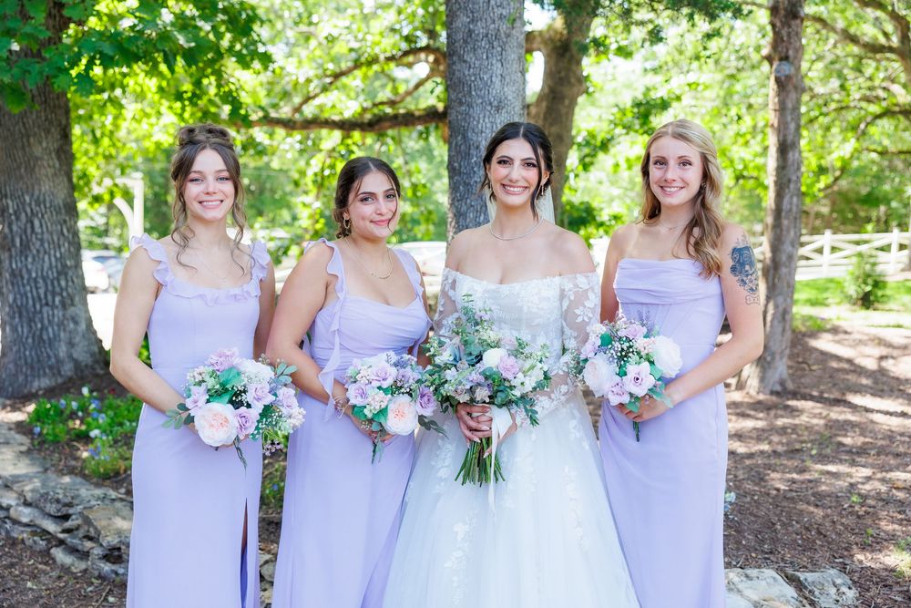 Four people in lavender bridesmaid dresses and a bride in white hold beautiful floral bouquets while standing outdoors.