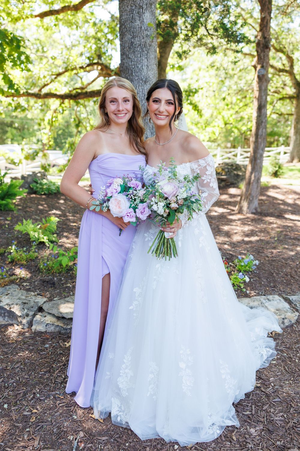 Two women in formal attire pose outdoors, one in a white wedding gown and one in a lavender bridesmaid dress.