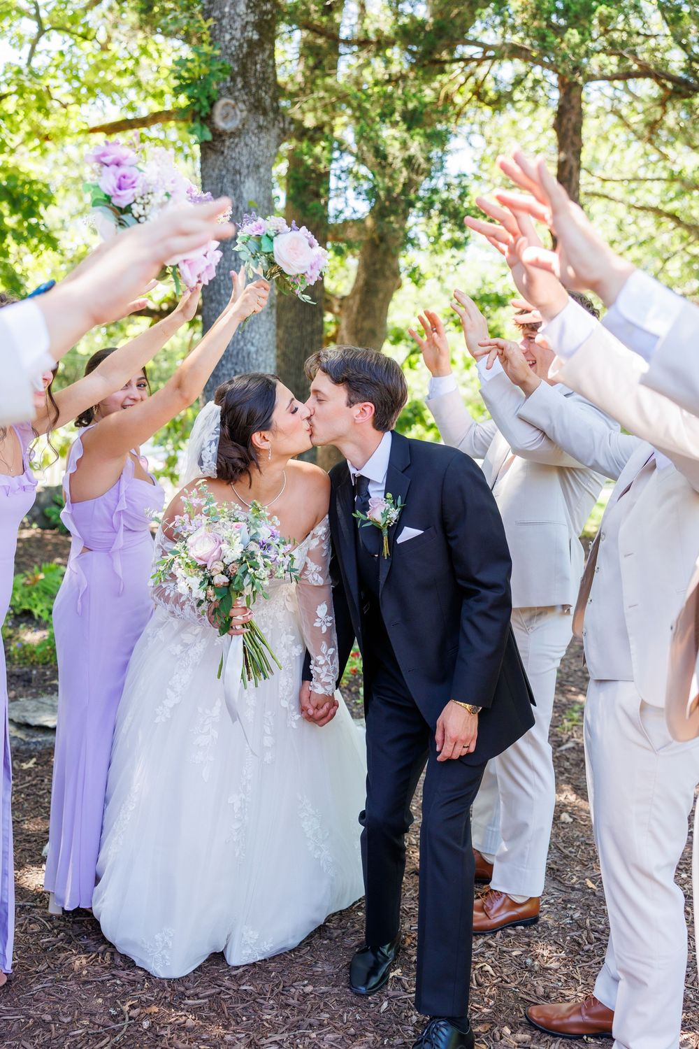 Wedding party celebrates with raised hands in an outdoor garden setting as the newlyweds share a kiss.