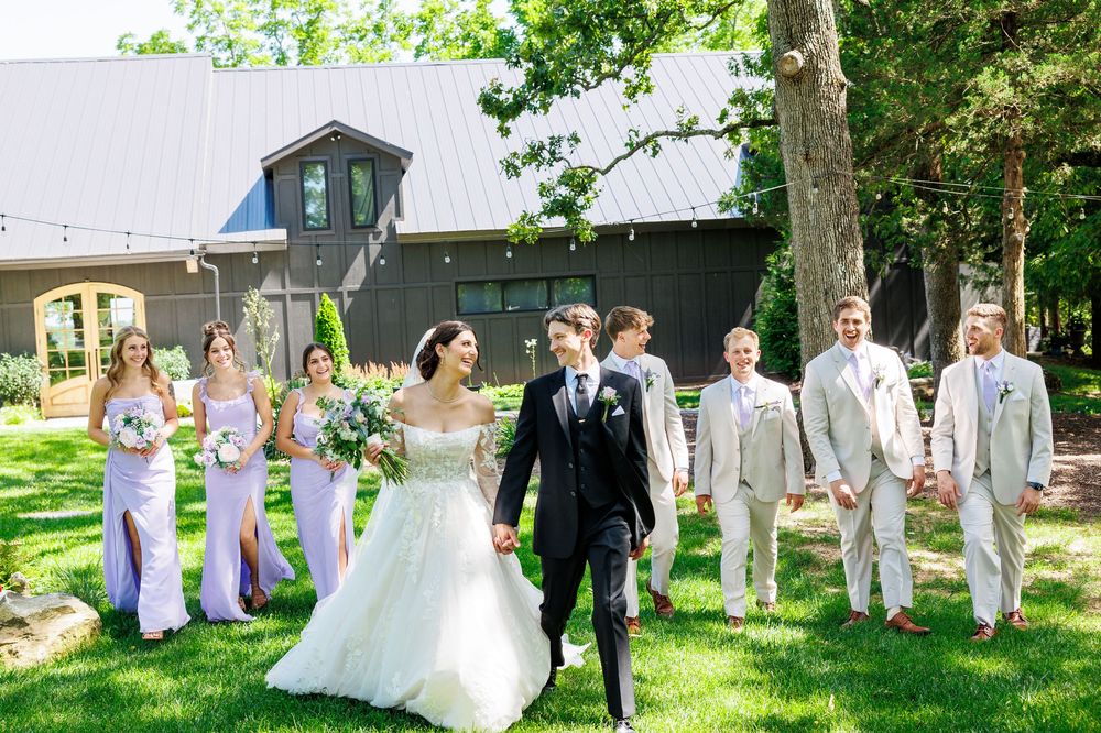 A wedding party gathers outside on a sunny summer day near a white building and lush green trees.
