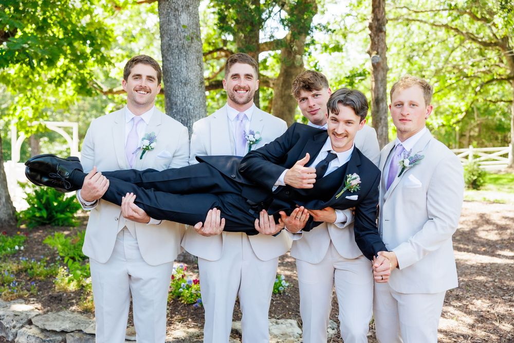 Groomsmen in light colored suits playfully lift someone during outdoor wedding photos.