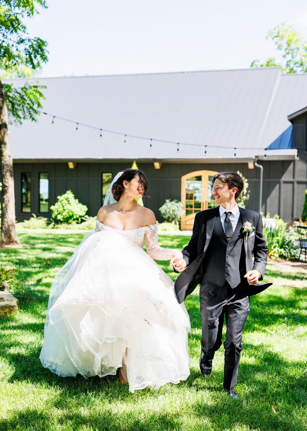 A couple runs playfully on grass near a tent decorated with string lights during their wedding celebration.