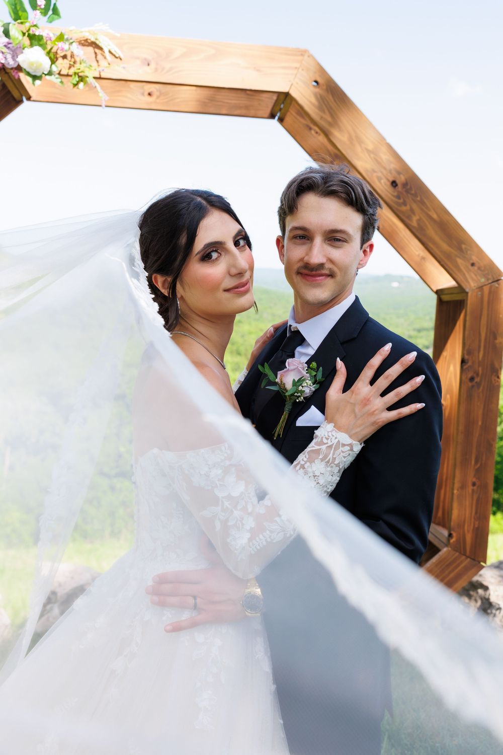 Romantic wedding couple poses under wooden hexagonal arch with flowing white veil in outdoor setting.