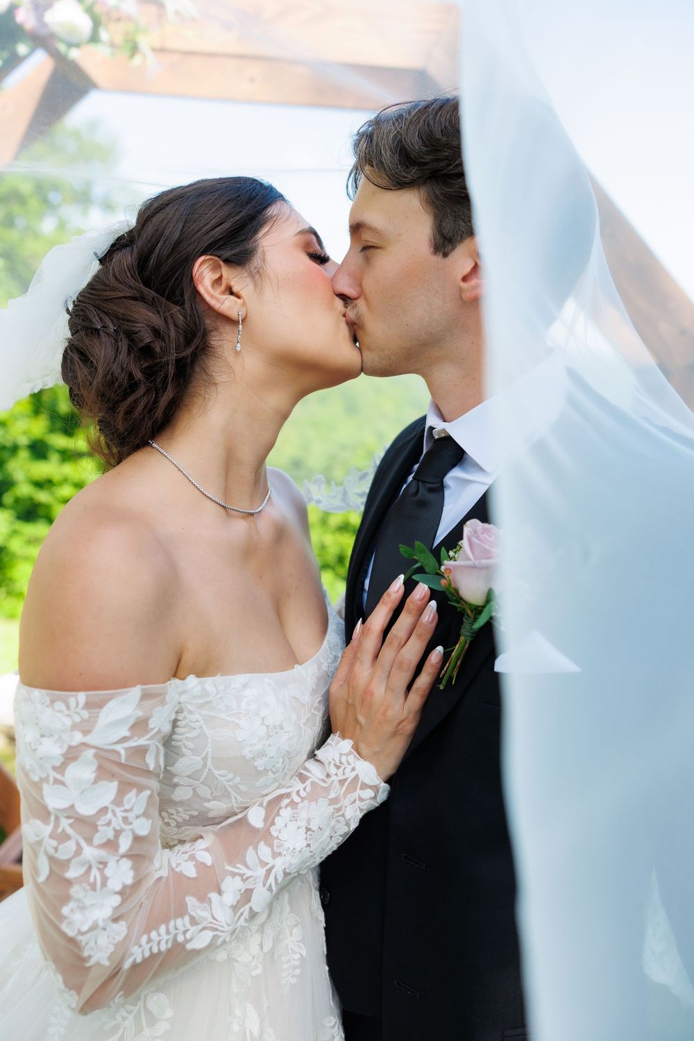 A romantic couple shares a tender kiss under flowing white veil during their outdoor wedding ceremony.
