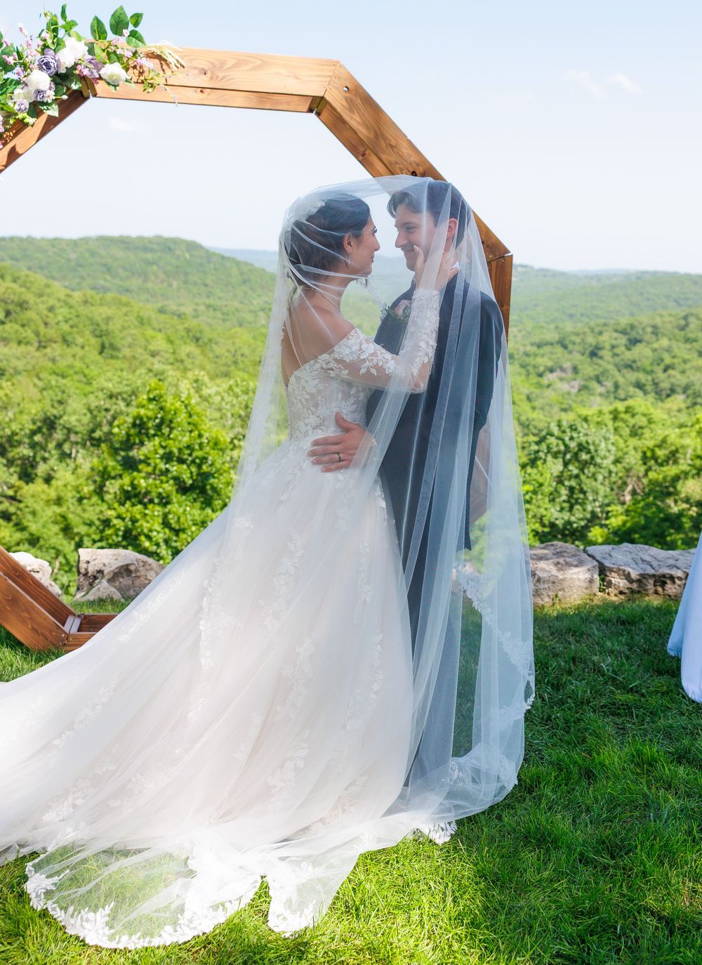 Wedding couple shares intimate moment under wooden arch against backdrop of rolling green hills.