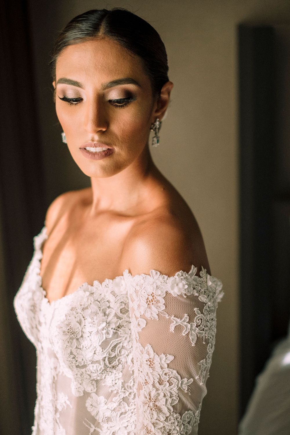 Close-up portrait of bride wearing an off-shoulder lace wedding gown and delicate earrings in soft lighting.