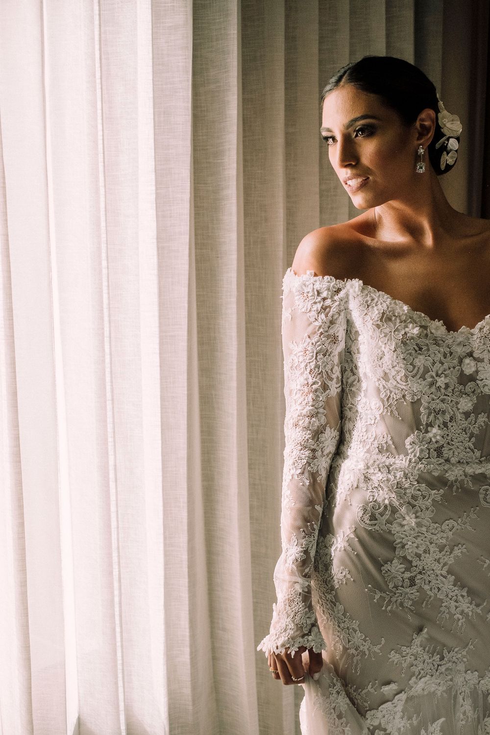 Elegant bride in off-shoulder lace wedding gown with long sleeves poses by window in soft natural light.