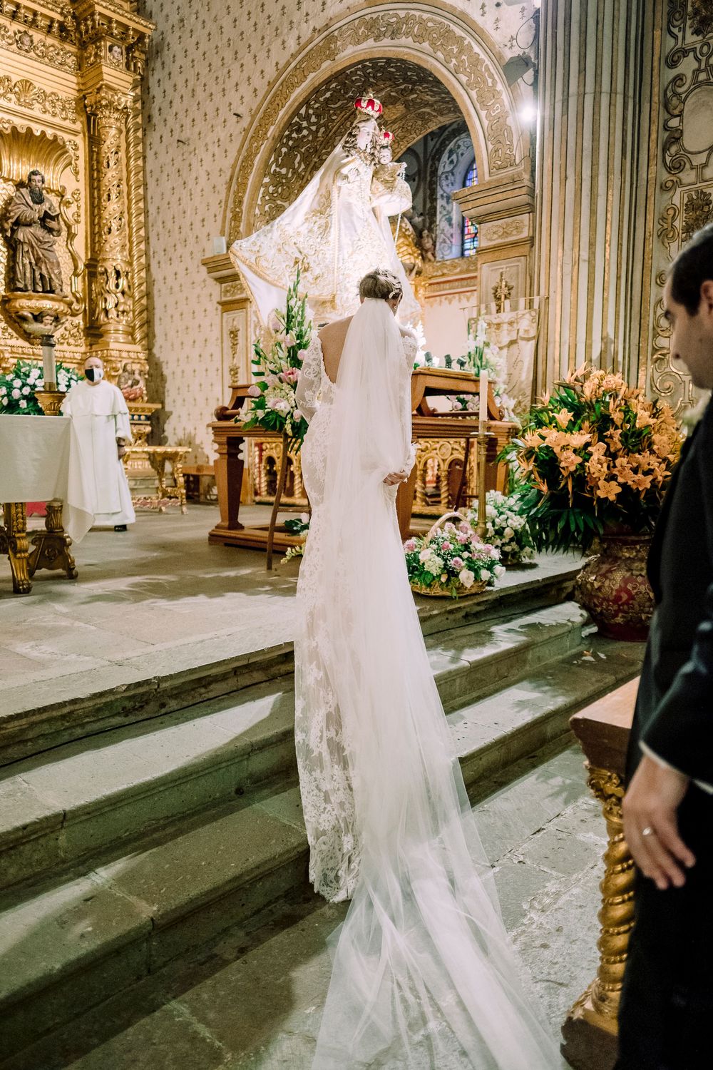 Bride in white lace gown and veil standing at ornate church altar with floral arrangements.