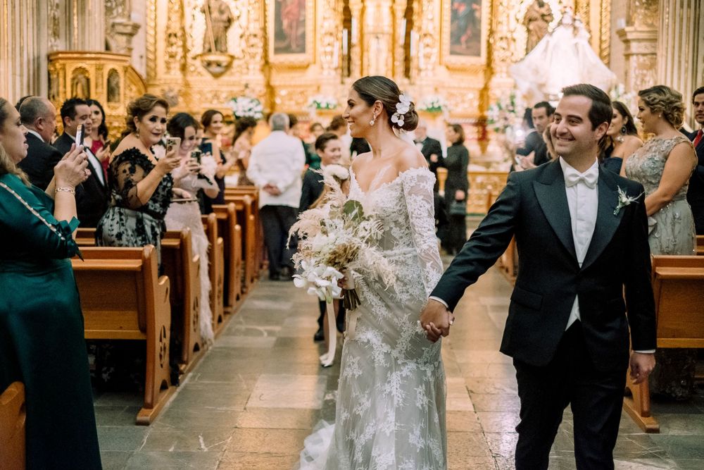 Wedding couple walks down church aisle while guests take photos in ornate cathedral interior.