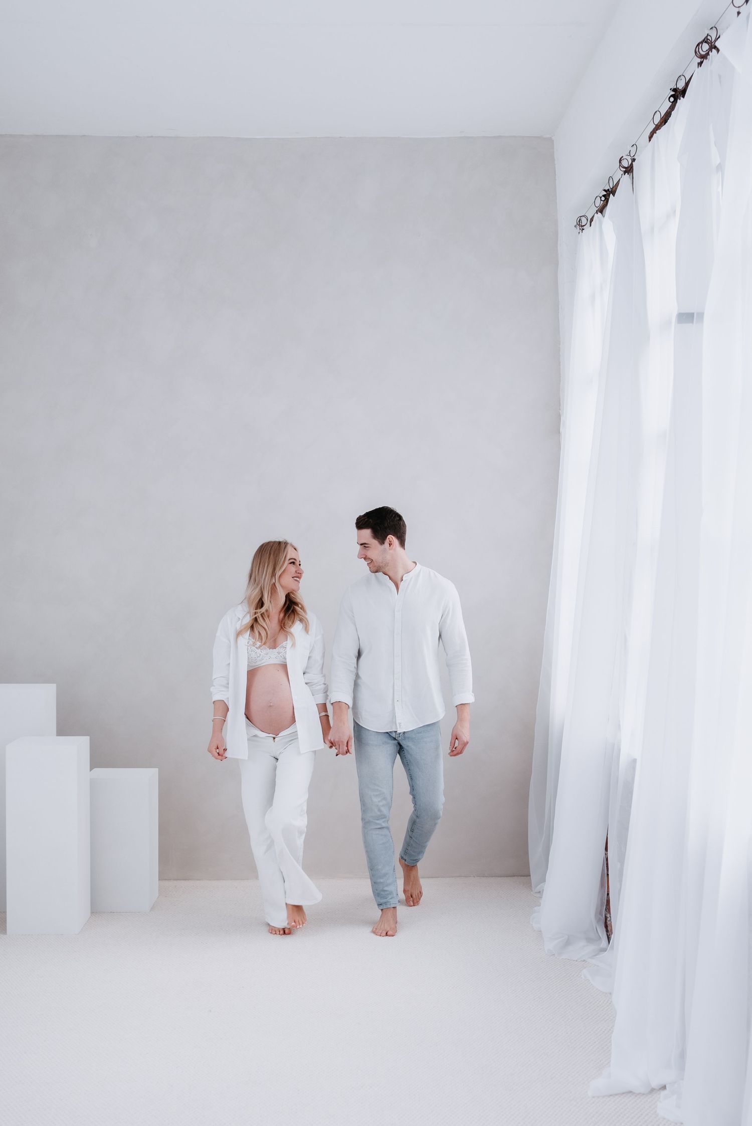 Couple in white outfits interact playfully in a bright minimalist studio space with white walls and floor.