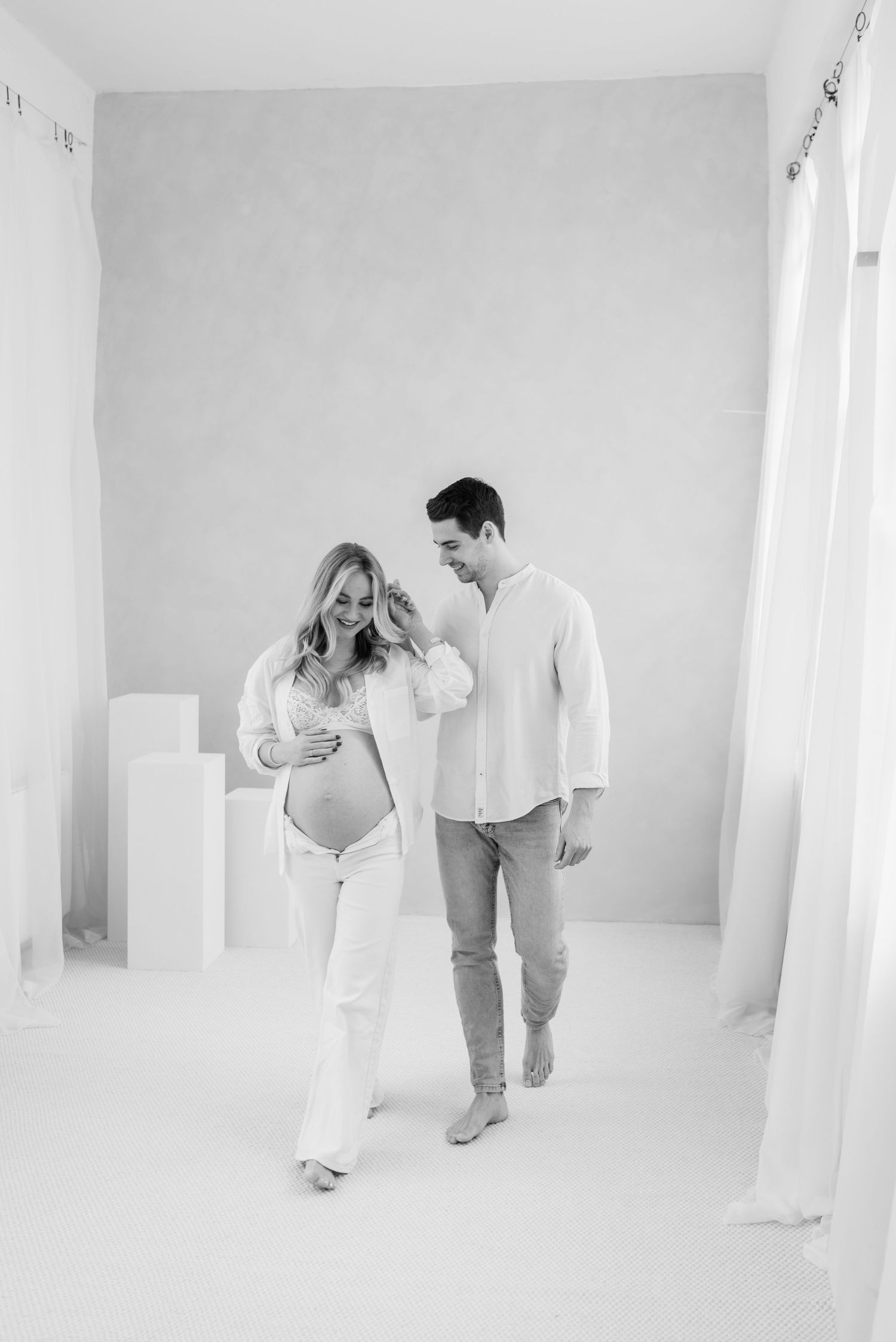 Black and white maternity photo of a couple walking together in an all-white studio setting with geometric shapes.