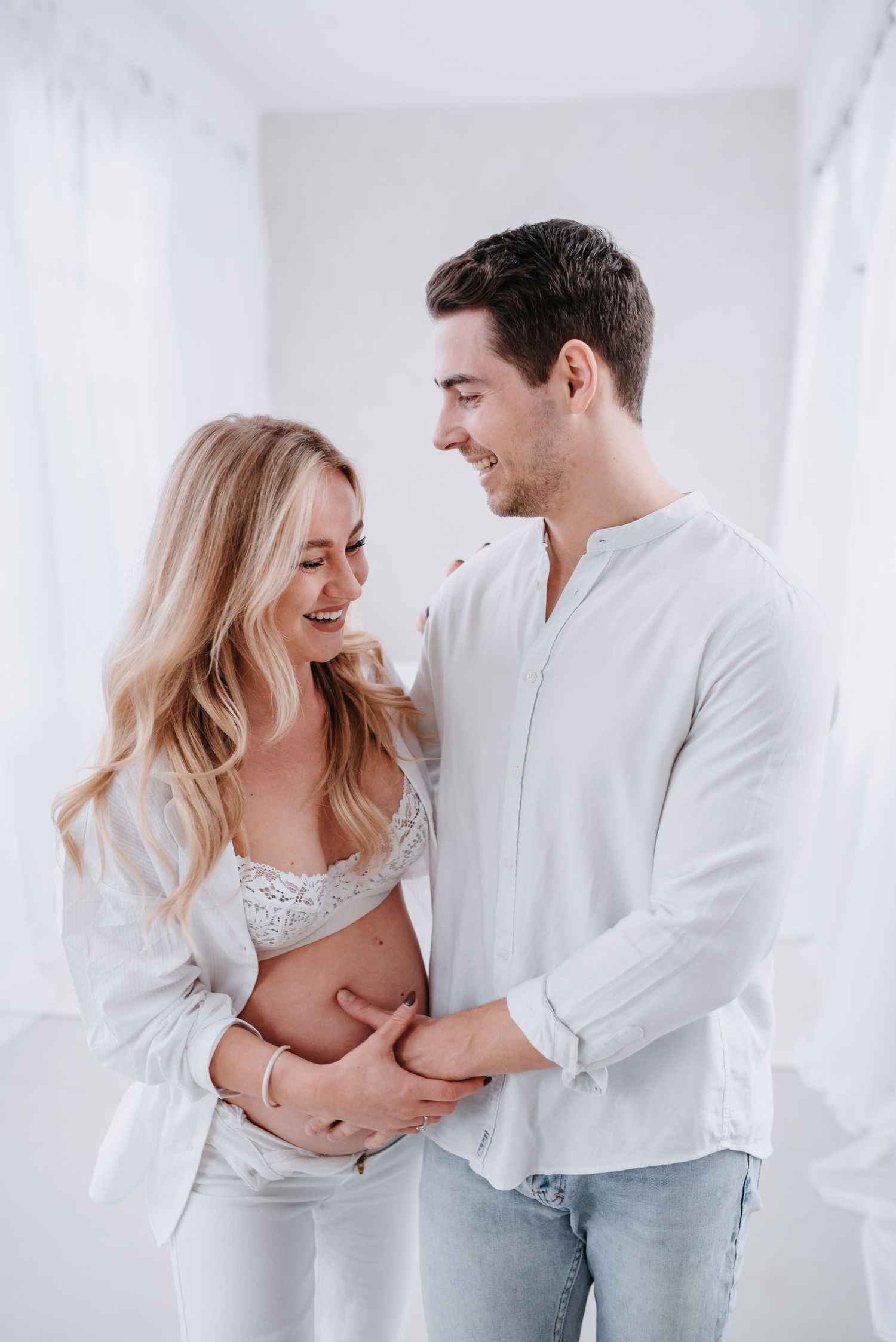 Joyful couple in white tops and light denim share intimate laughs in a bright minimalist setting.