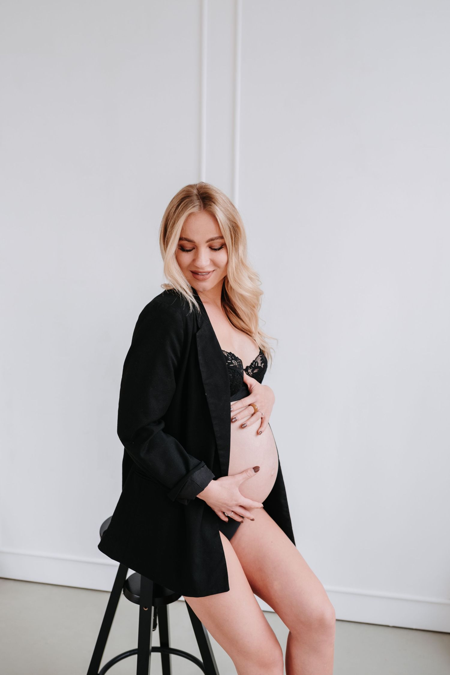 A pregnant woman wearing black lingerie and an open cardigan poses on a stool against a white wall.