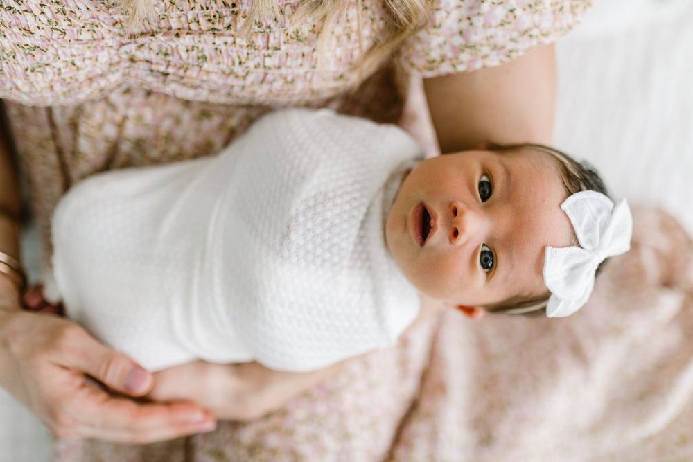 Newborn baby in white wrap and bow looks up while resting on soft pink fabric.