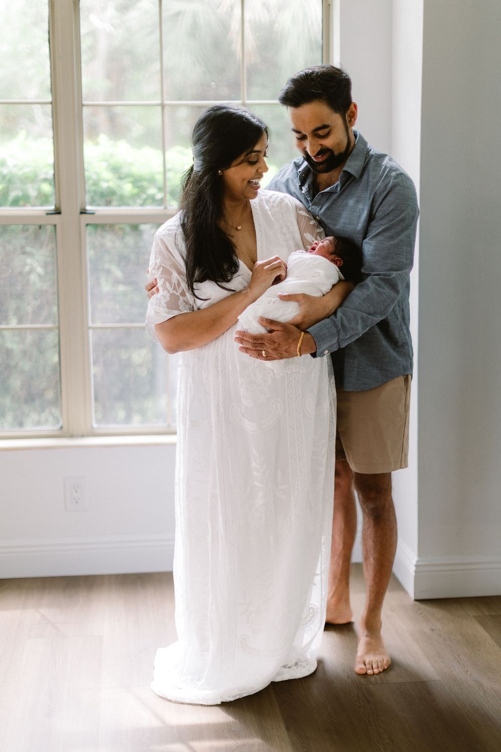 New parents in casual attire holding their swaddled baby by bright window.