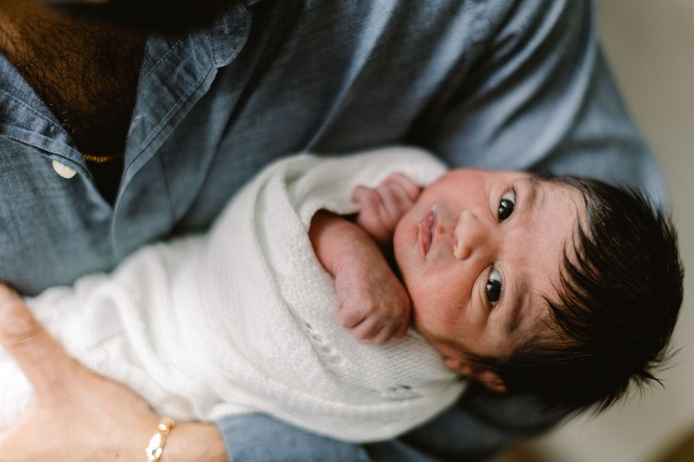 Alert newborn wrapped in white swaddle being held against gray shirt.