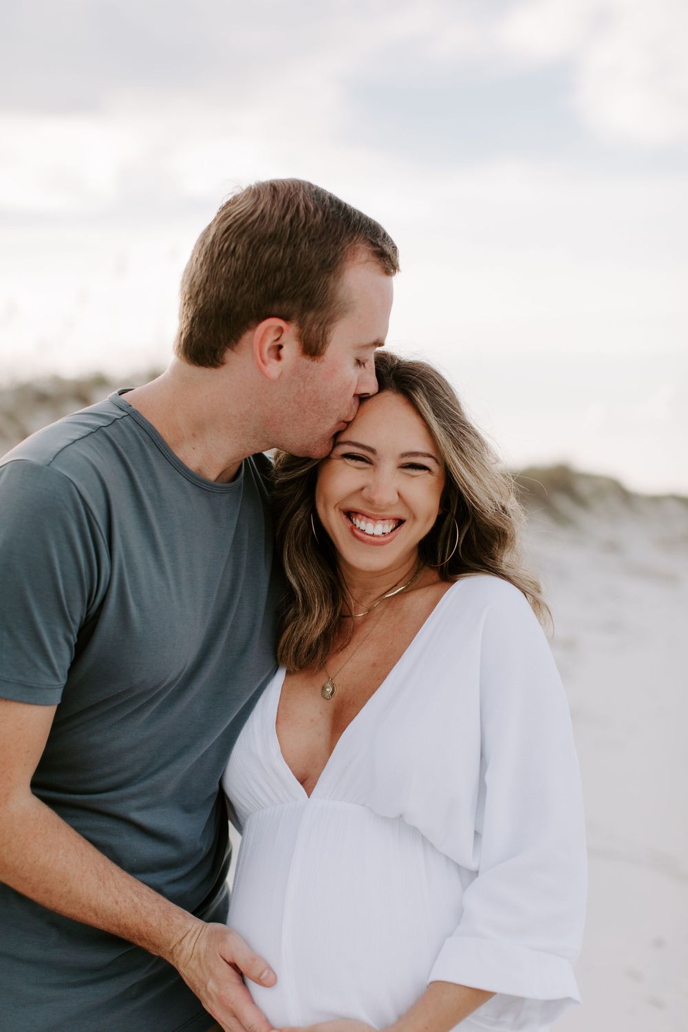 A romantic maternity photoshoot showing a couple embracing on sandy dunes while wearing casual clothing.