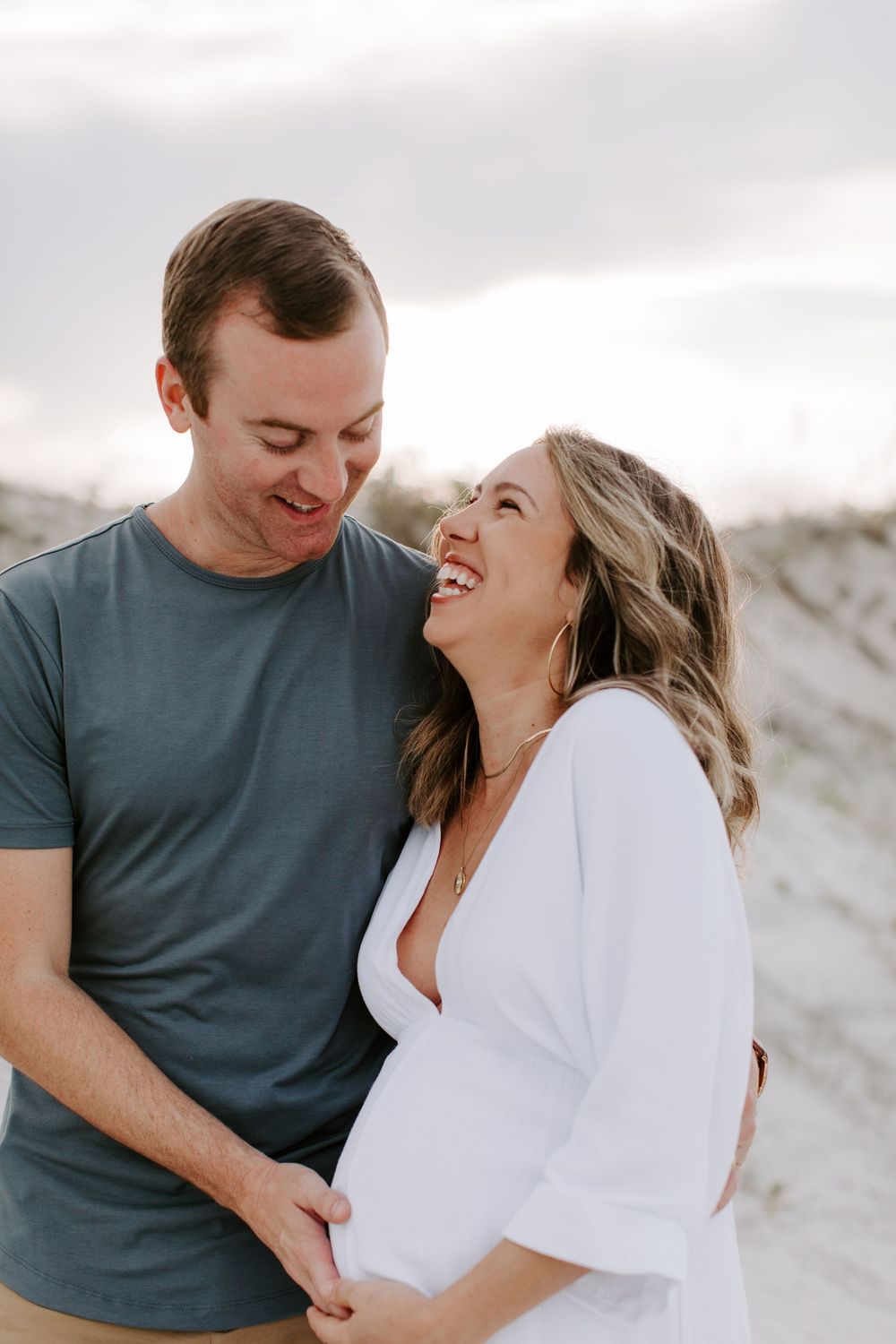Candid moment of a couple laughing together during their maternity photo session on a beach.