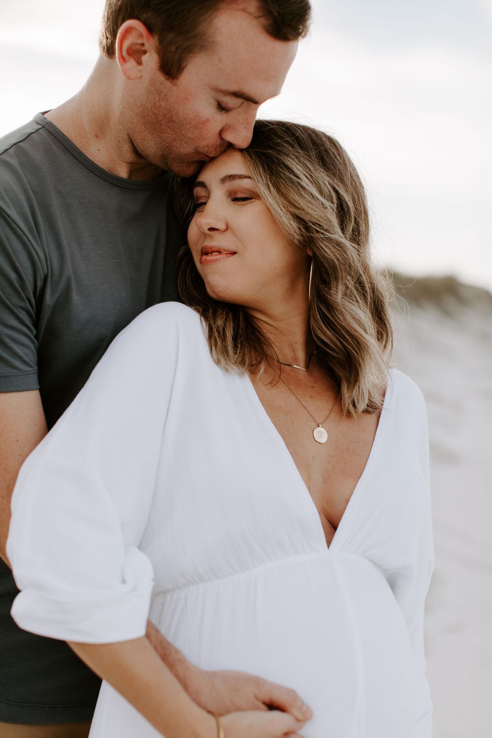 Intimate maternity portrait of a couple in white and grey attire against sand dunes.