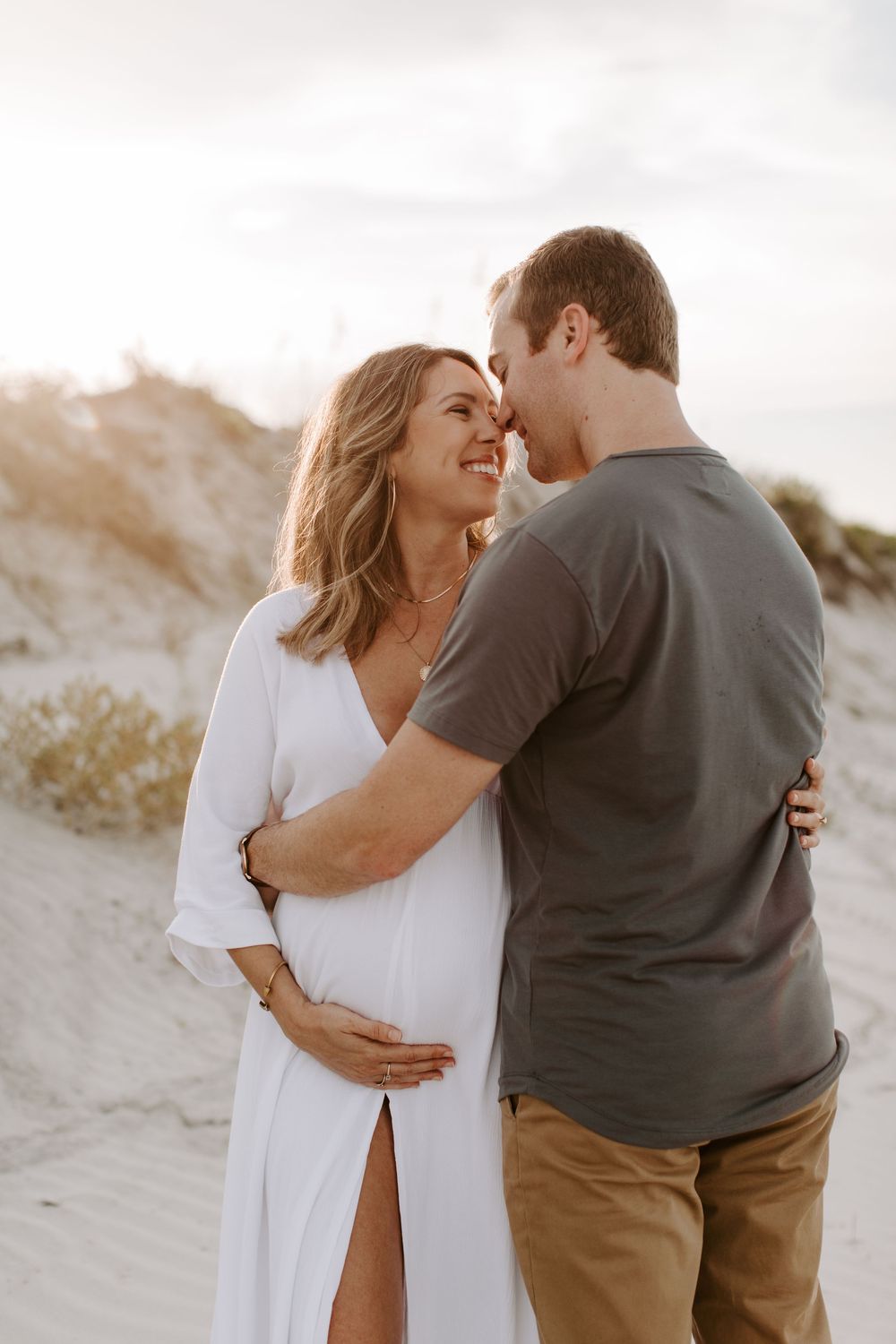 Loving couple in casual attire sharing tender moment during beach maternity photoshoot.