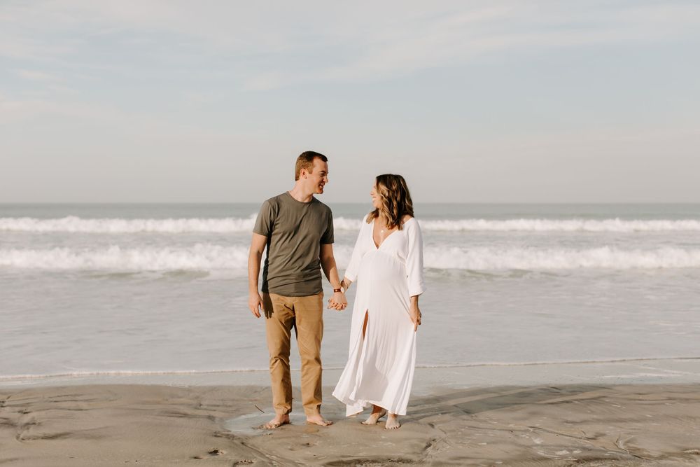 A couple holding hands while walking along the shoreline during their maternity photoshoot.
