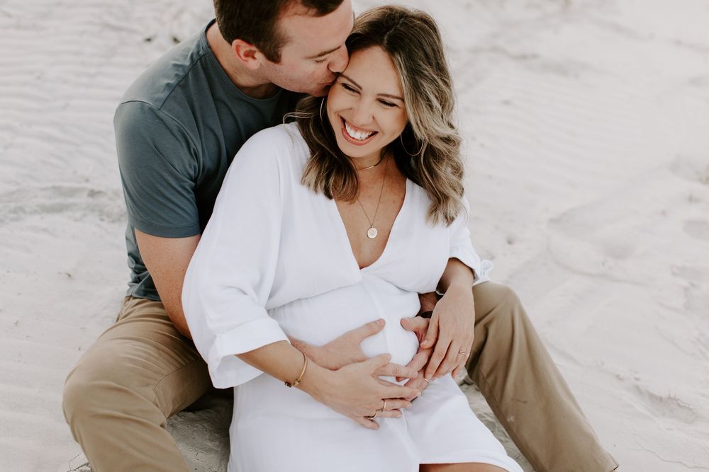 Sweet beach maternity photo of couple sitting on sand, sharing intimate moment.